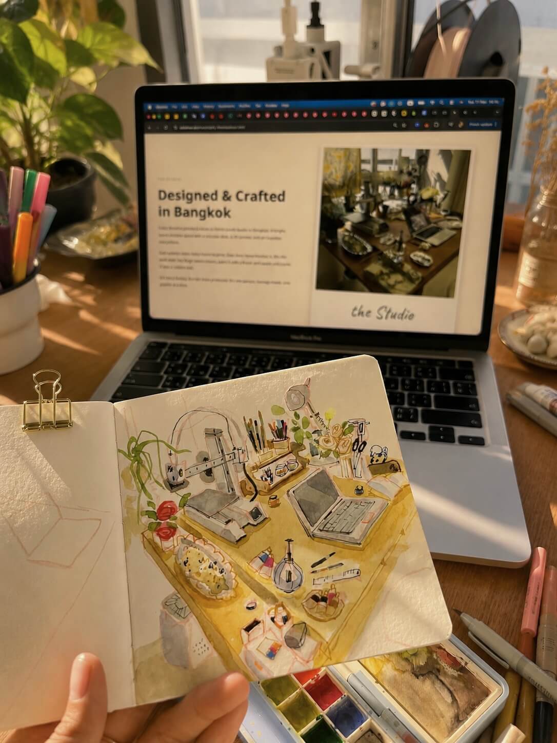 Watercolour sketchbook illustration held in front of studio MacBook